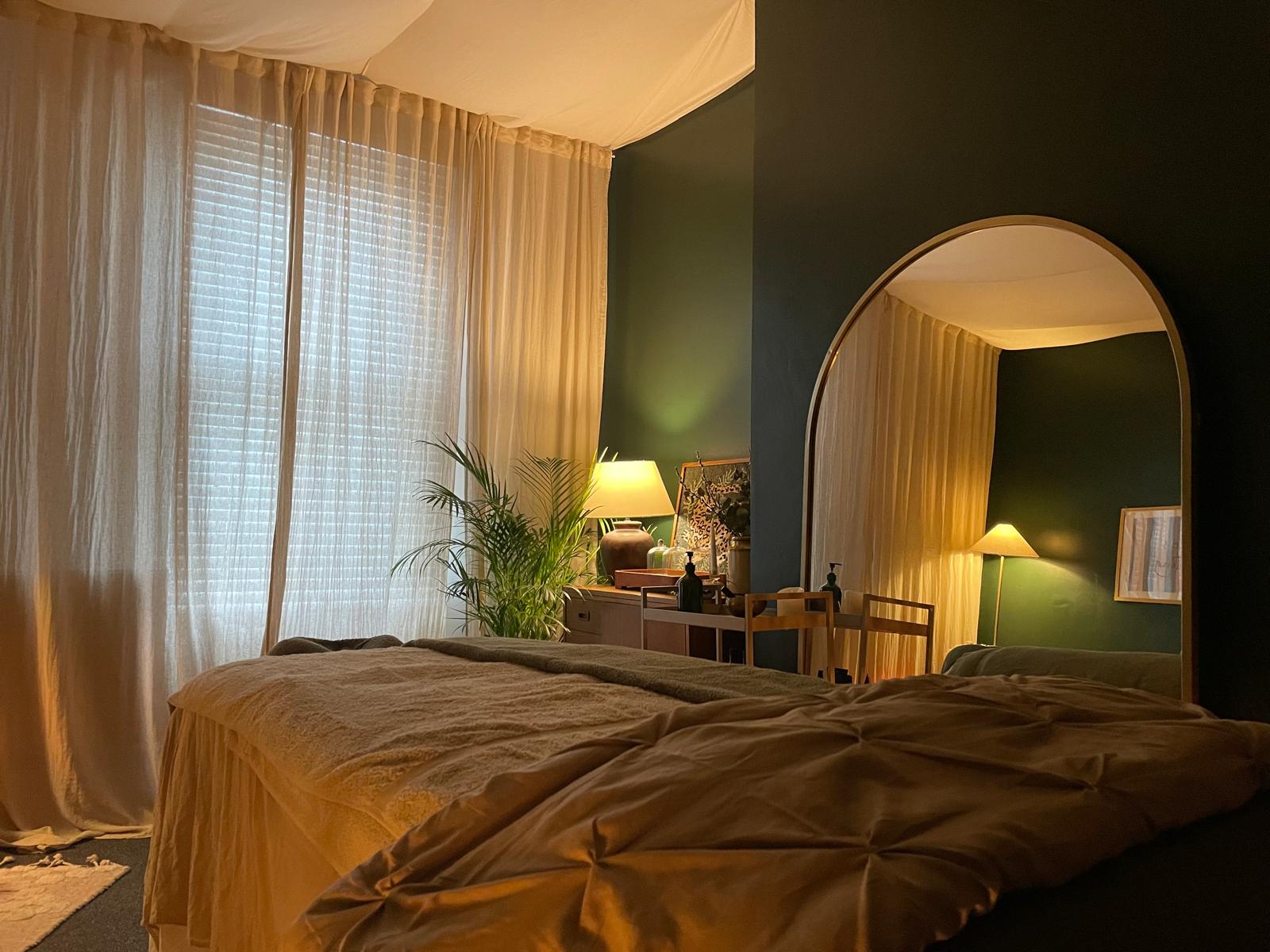 Bee's treatment room at dusk: sheer curtains, arched mirror, warm lamplight on the massage table.
